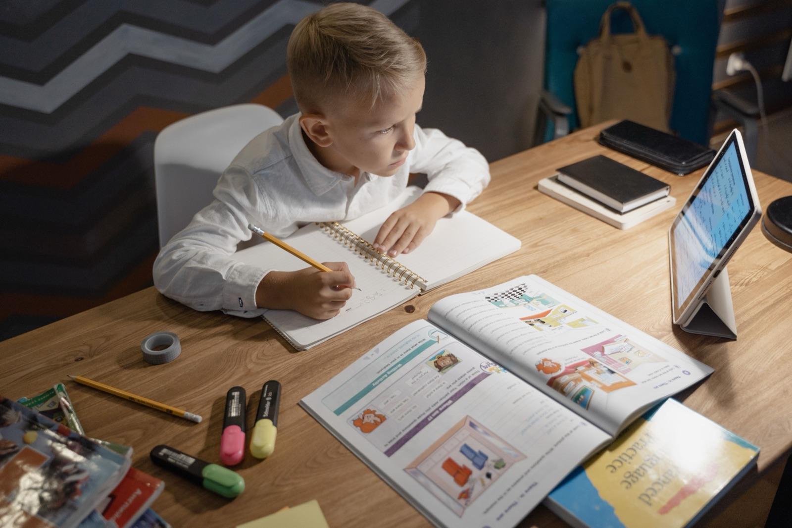 A middle-school-age boy working through a maths workbook beside a tablet