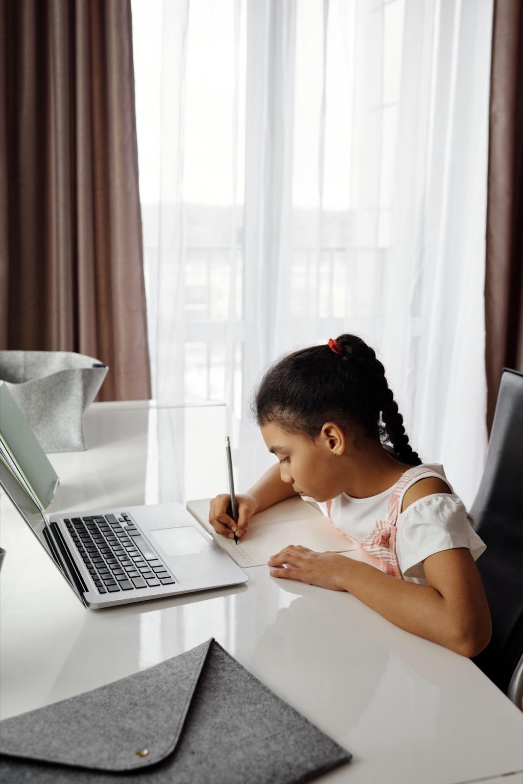 A child working through a lesson at a laptop, writing notes by hand